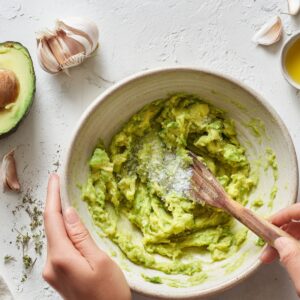 Hands holding a bowl of mashed avocado, with a wooden spoon mixing in salt. Around the bowl are garlic cloves, olive oil, and a small wooden bowl of more salt.