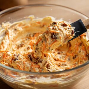 Mixing bowl of carrot cake batter with shredded carrots, pecans, and creamy mixture being stirred with a spatula.