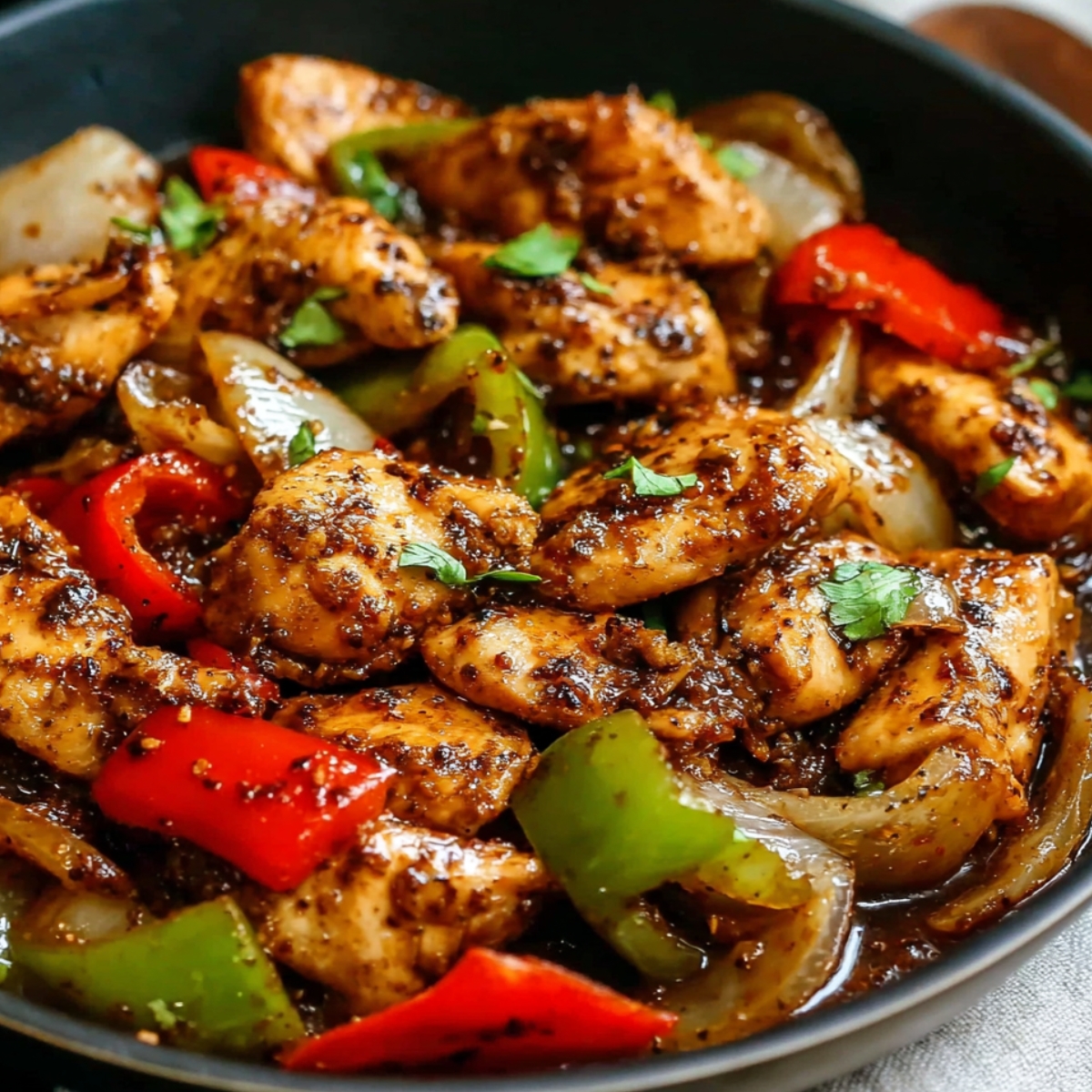 Black pepper chicken stir-fry with tender chicken chunks, red and green bell peppers, onions, and fresh cilantro garnish in a dark bowl.