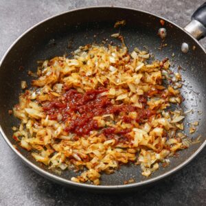 Onions sautéing in a pan until golden brown, with a dollop of rich red tomato paste added to the center for cooking.