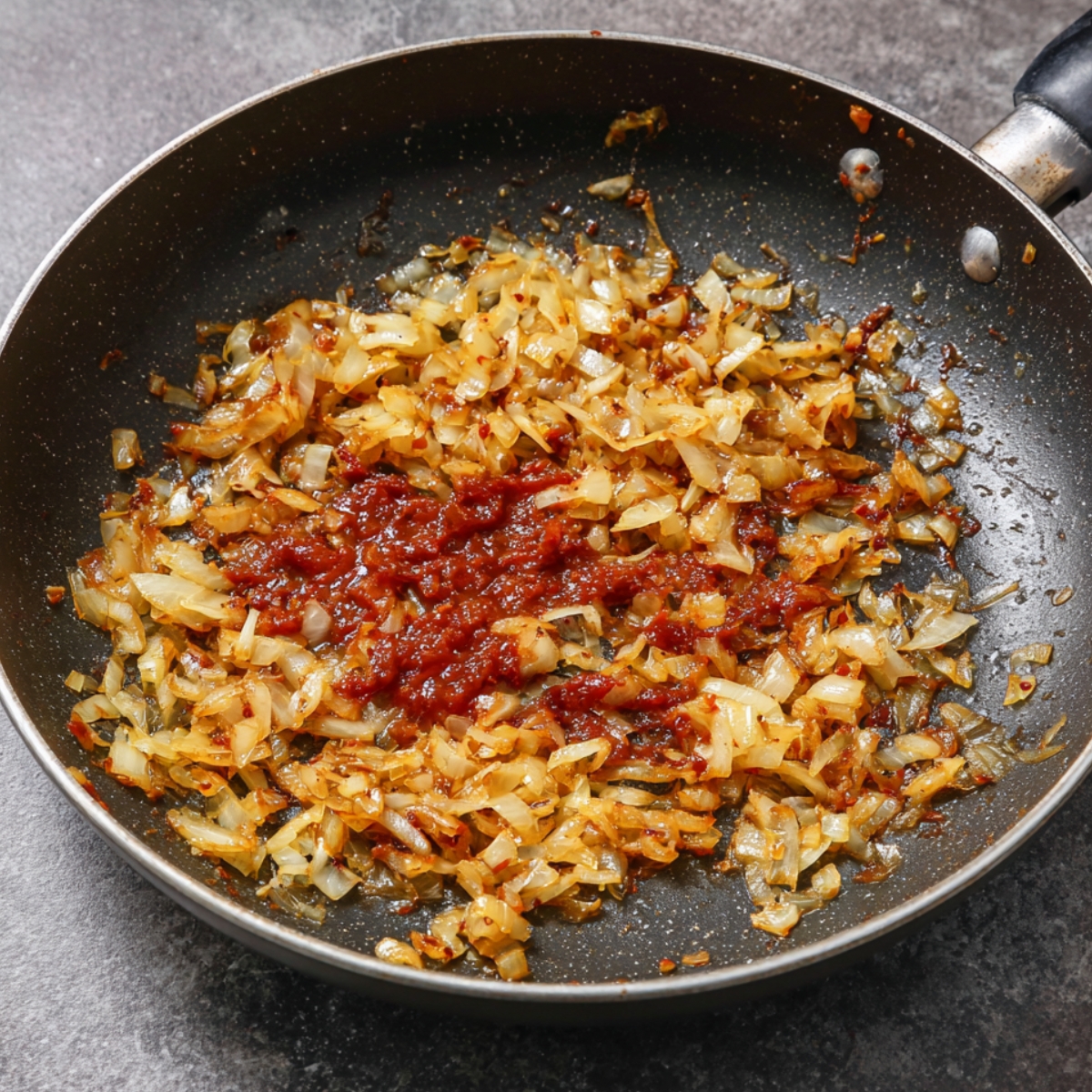 Onions sautéing in a pan until golden brown, with a dollop of rich red tomato paste added to the center for cooking.