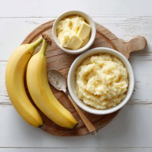 Two ripe bananas on a wooden board beside a bowl of mashed banana mixture and another small bowl filled with butter, with a spoon resting nearby.