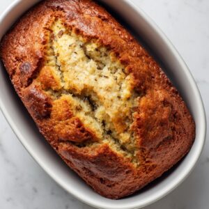 A freshly baked golden-brown loaf of banana bread in a white baking dish, with a cracked top showing its soft, moist texture inside.