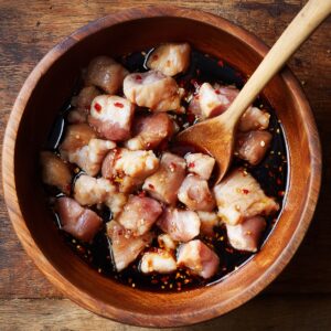 Raw chicken pieces marinating in soy sauce with garlic and chili flakes in a wooden bowl, stirred with a wooden spoon.