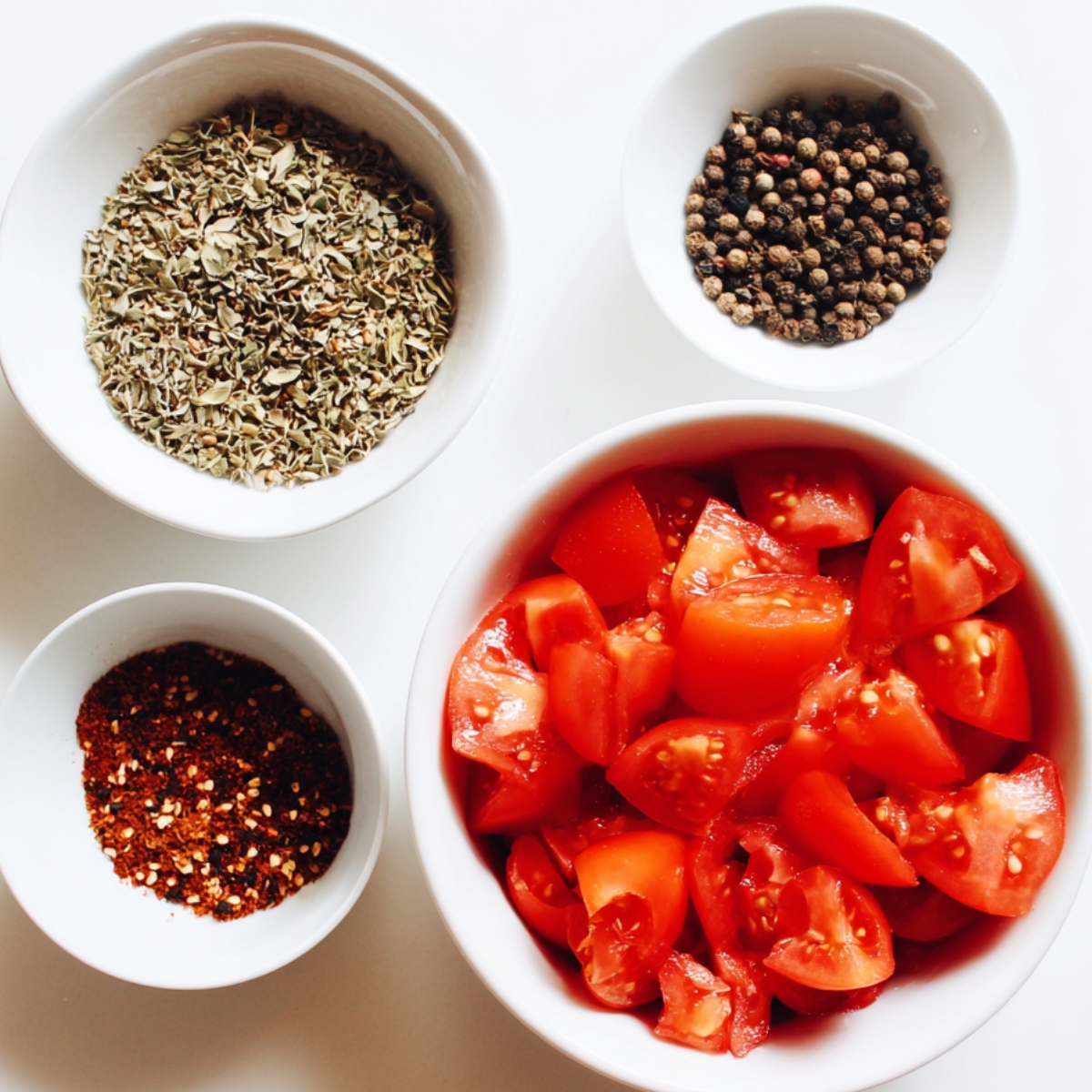 Four small white bowls on a white background. One contains chopped fresh tomatoes, another has whole black peppercorns, another holds dried oregano, and the last one has red chili flakes.