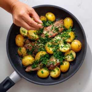 "A hand sprinkling fresh chopped herbs over sizzling steak slices and baby potatoes in a frying pan."