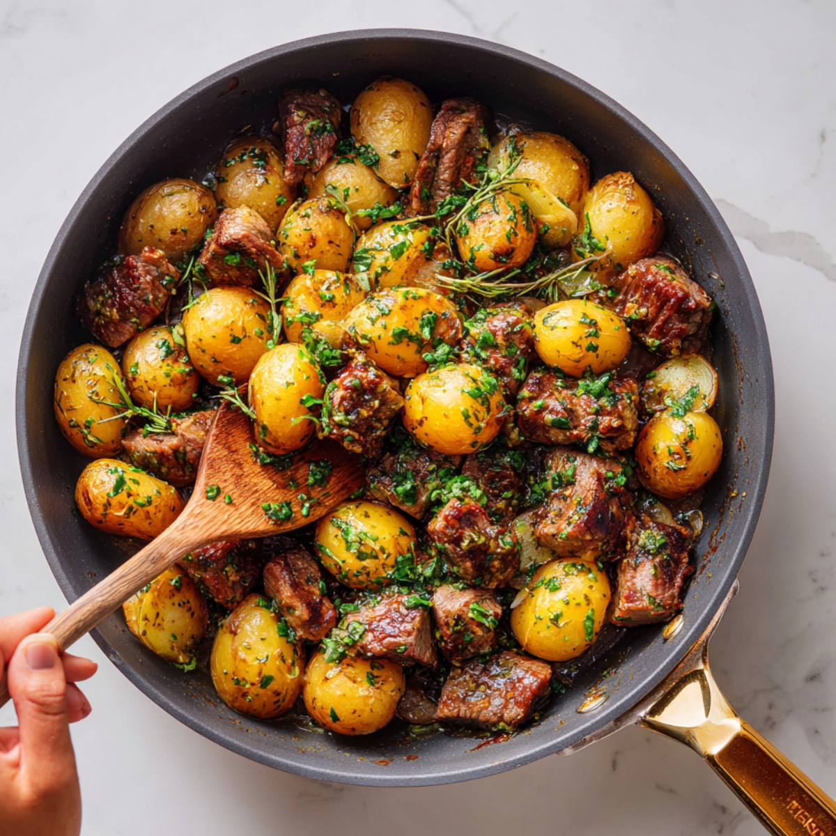 "A skillet filled with golden baby potatoes and seared steak bites, garnished with fresh herbs and stirred with a wooden spoon."