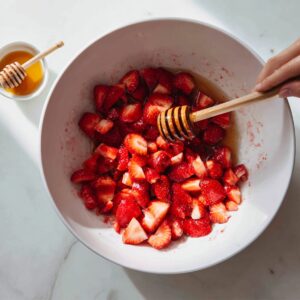 A bowl filled with freshly cut strawberries being mixed with honey using a wooden honey dipper, with vibrant red strawberries glistening under the light.