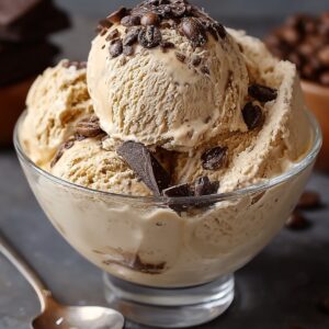 A glass bowl filled with scoops of creamy Homemade Coffee Ice Cream Recipe, topped with coffee beans and chunks of dark chocolate, placed on a dark surface with a spoon beside it.
