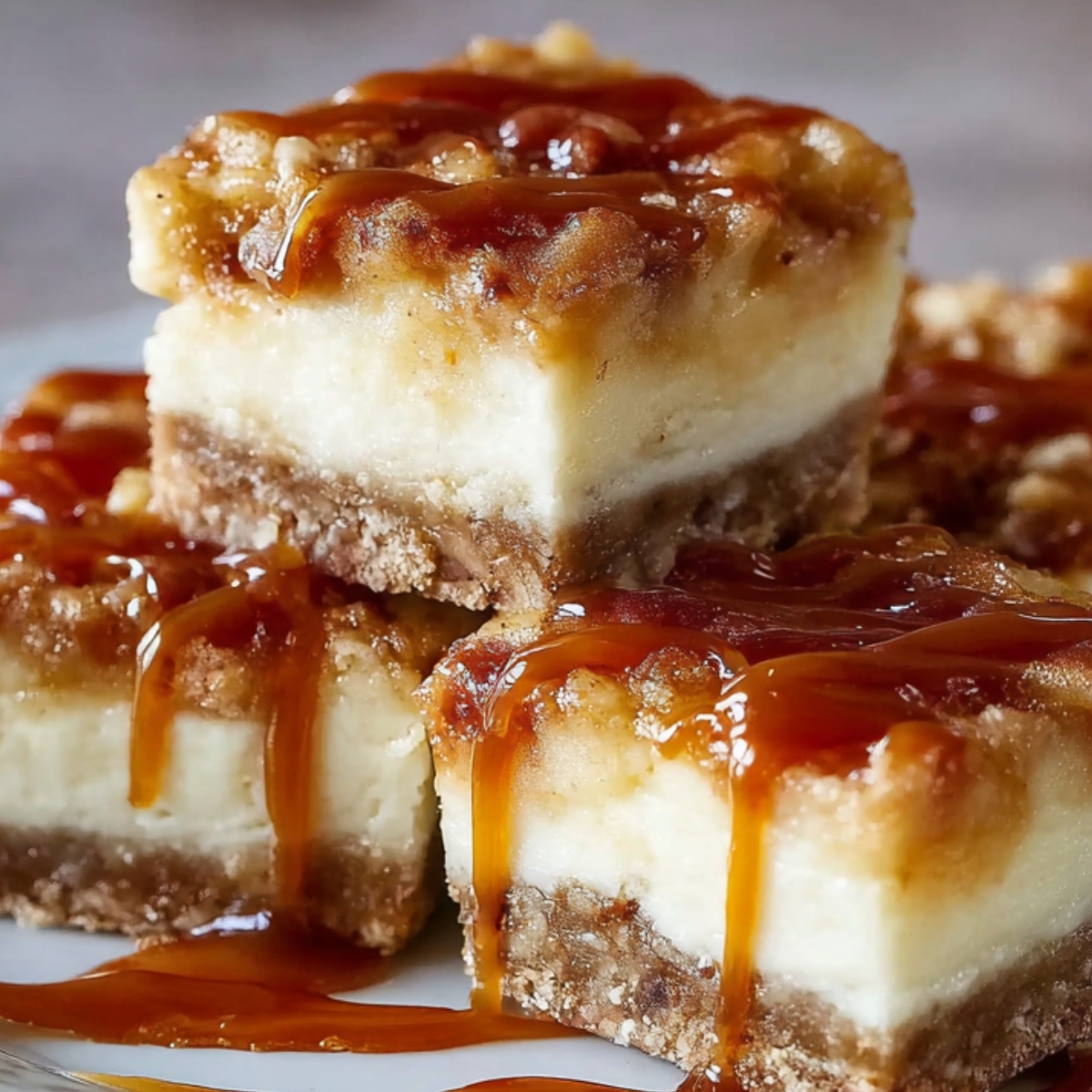 Homemade caramel apple cheesecake bars stacked on a plate and drizzled with golden caramel sauce.