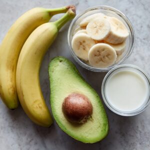 Raw smoothie ingredients arranged on a countertop: fresh bananas, a halved avocado, a small bowl of milk, and sliced banana in a glass bowl.