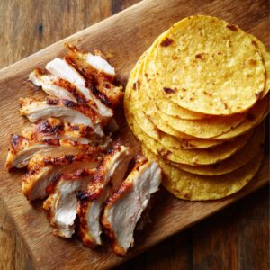 Sliced grilled chicken breast arranged beside a neat stack of yellow corn tortillas on a wooden cutting board.