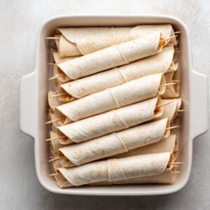 A white baking dish filled with uncooked, rolled chicken flautas secured with toothpicks, made with flour tortillas.