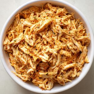 A bowl of shredded chicken seasoned with spices and herbs, ready to be used as filling for dishes like flautas or tacos.