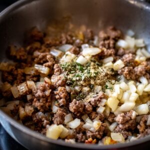 Ground beef and onions cooking in a skillet with herbs and spices sprinkled on top.