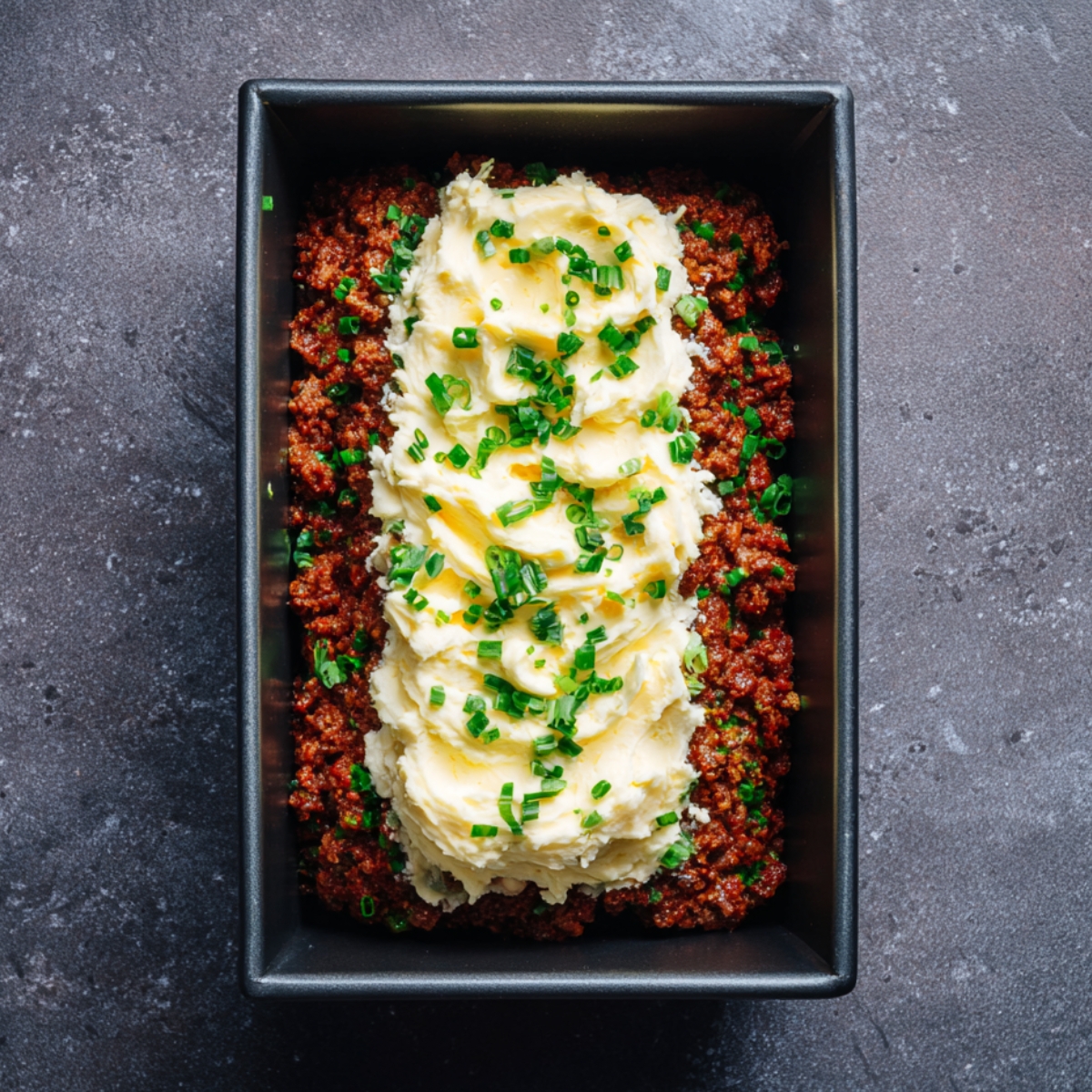 A rectangular pan filled with jalapeño popper meatloaf topped with mashed potatoes, melted butter, and chopped green onions. The meat is finely ground and reddish from seasoning.