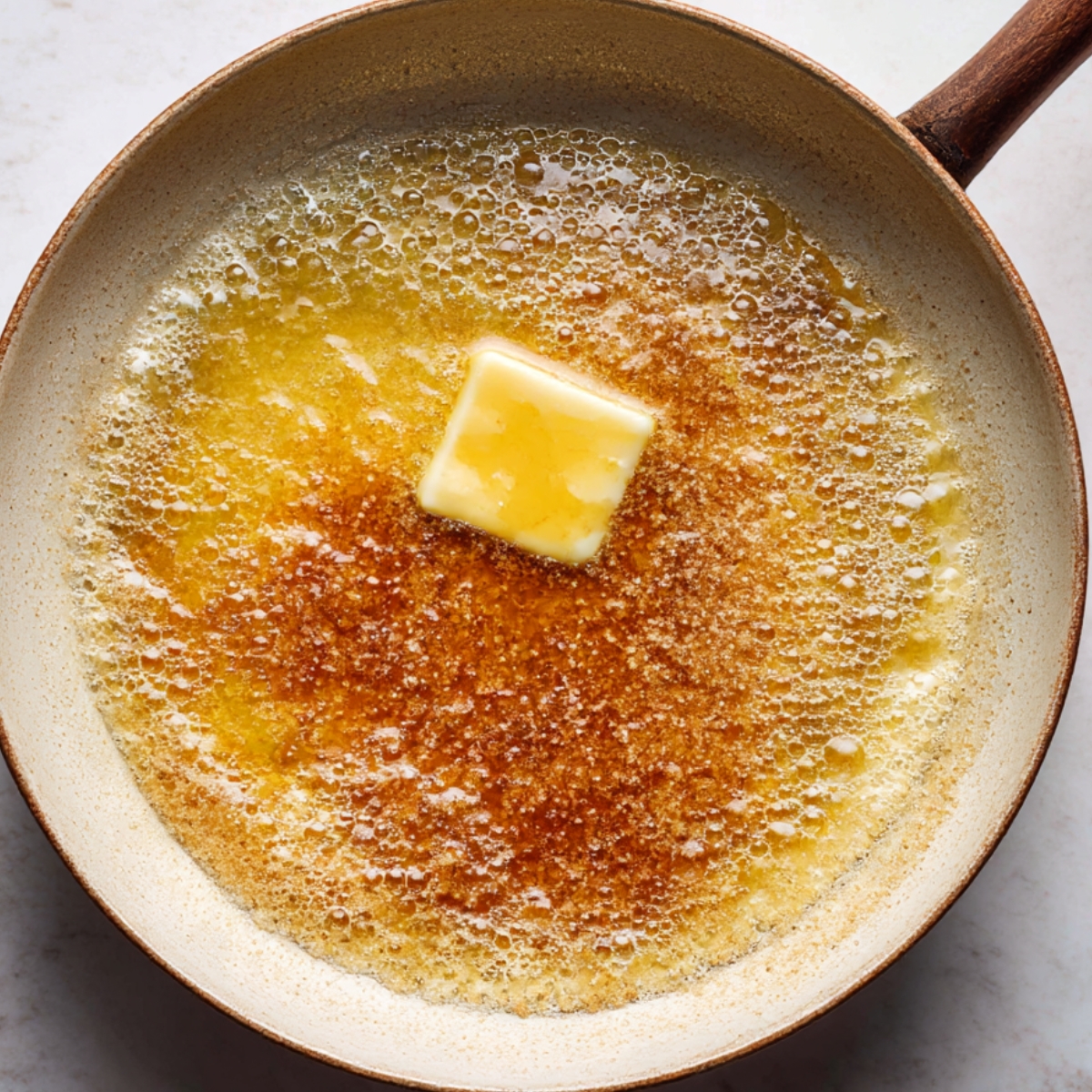 A close-up of a skillet with butter melting in a golden pool of bubbling liquid, with a slight caramelization effect around the edges, suggesting it's the start of the process for preparing the topping for a Pineapple Upside Down Cake.