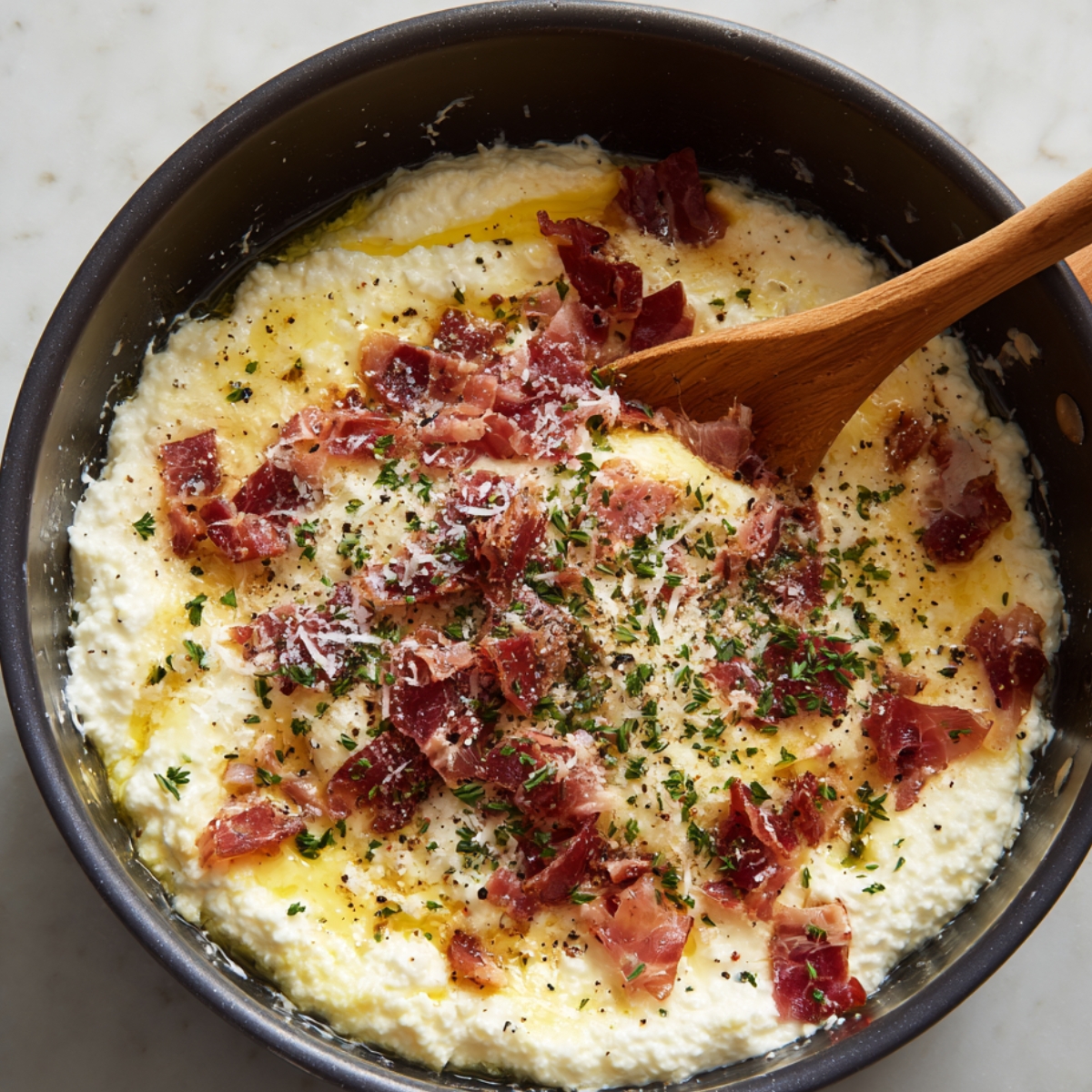 A black pan with creamy ricotta mixture topped with chopped cured meat, grated cheese, herbs, and black pepper, being stirred with a wooden spoon.