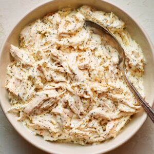 Bowl of shredded chicken mixed with creamy sauce and poppy seeds, with a spoon resting inside.