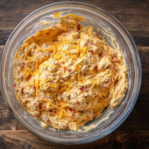 A large glass mixing bowl filled with uncooked sausage ball mixture, showing visible bits of cheddar cheese, sausage, and diced Rotel tomatoes in a creamy base.