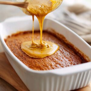 A golden, glossy toffee sauce being poured over a freshly baked sticky toffee pudding in a white rectangular baking dish.