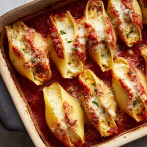 A close-up shot of a baking dish with baked stuffed manicotti shells, topped with tomato sauce, melted cheese, and fresh herbs.