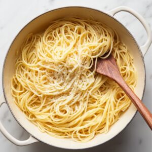 Cooked spaghetti pasta in a large pot, tossed with grated parmesan cheese and a wooden spoon.