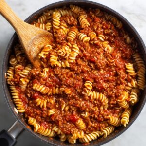 A pan full of rotini pasta mixed with ground beef and tomato meat sauce, being stirred with a wooden spoon.