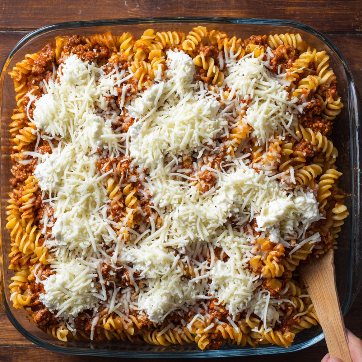 A glass baking dish filled with layered rotini pasta, meat sauce, ricotta cheese, and shredded mozzarella cheese, ready to be baked.