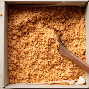 Crushed graham cracker crust mixture being spread evenly in a square baking pan.