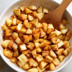 Chopped apples mixed with cinnamon and sugar in a white bowl with a wooden spoon.