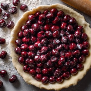 Unbaked cherry pie with a deep red cherry filling generously piled into a raw pie crust, surrounded by fresh cherries and flour.