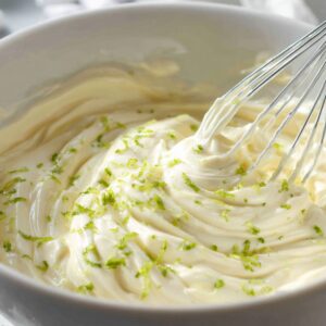Whisk stirring creamy key lime mixture with flecks of lime zest in a white bowl.