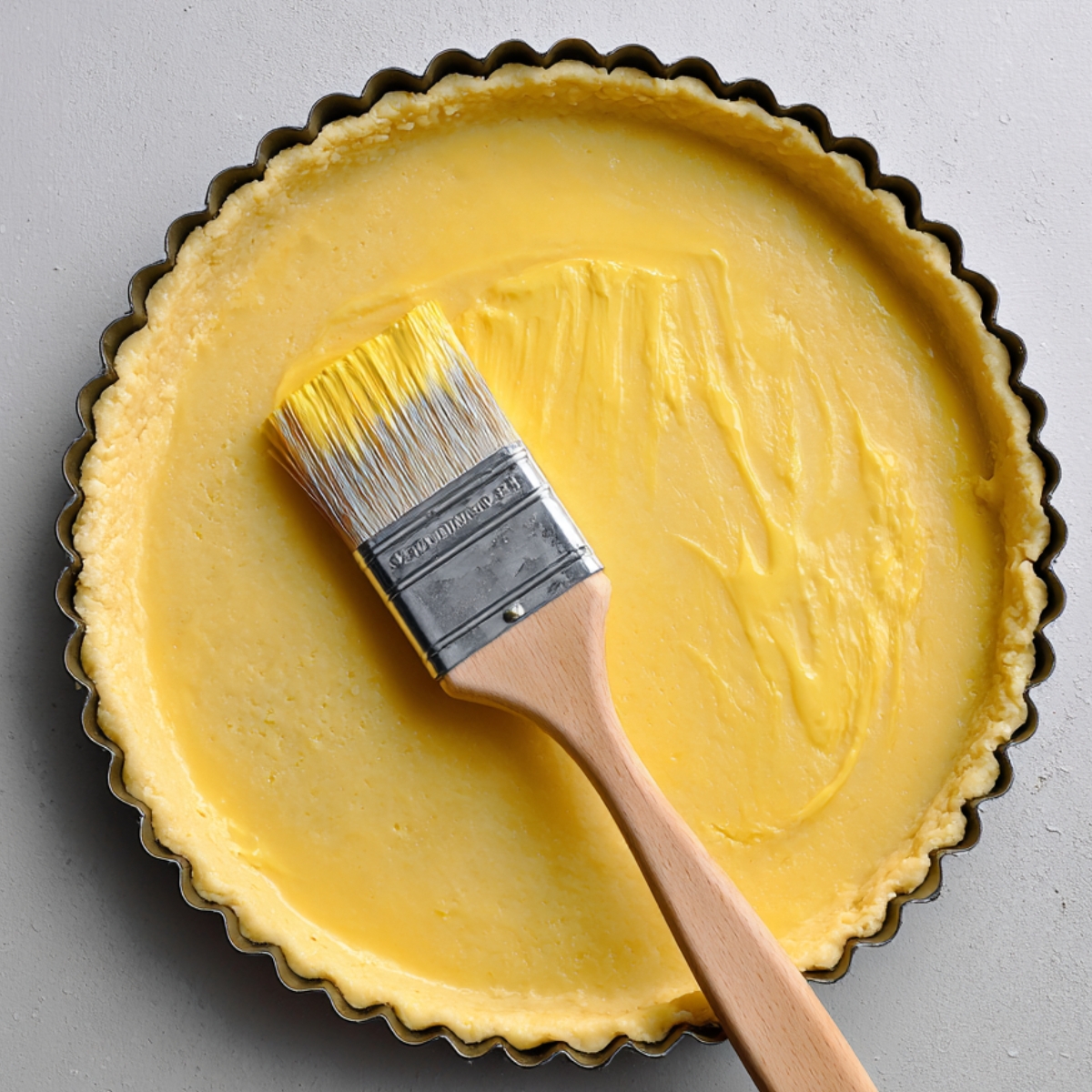 A raw tart shell being brushed with an egg wash using a pastry brush inside a round tart pan.