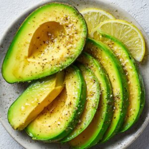 Sliced avocado arranged on a plate with cracked black pepper, sea salt, and lemon wedges.