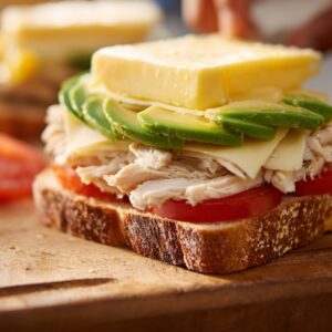 Open-faced sandwich on a wooden board being prepared with layers of sliced tomato, shredded chicken, avocado slices, and cheese.