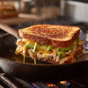 Chicken avocado melt sandwich grilling in a cast-iron skillet, with golden brown crispy bread and melted cheddar cheese dripping down.