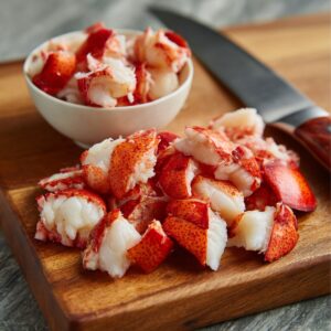 Chopped lobster meat on a wooden cutting board next to a white bowl and a knife, ready for making lobster salad.