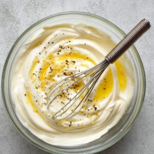 Glass bowl of homemade dressing with mayonnaise, lemon zest, olive oil, and cracked black pepper, with a metal whisk.