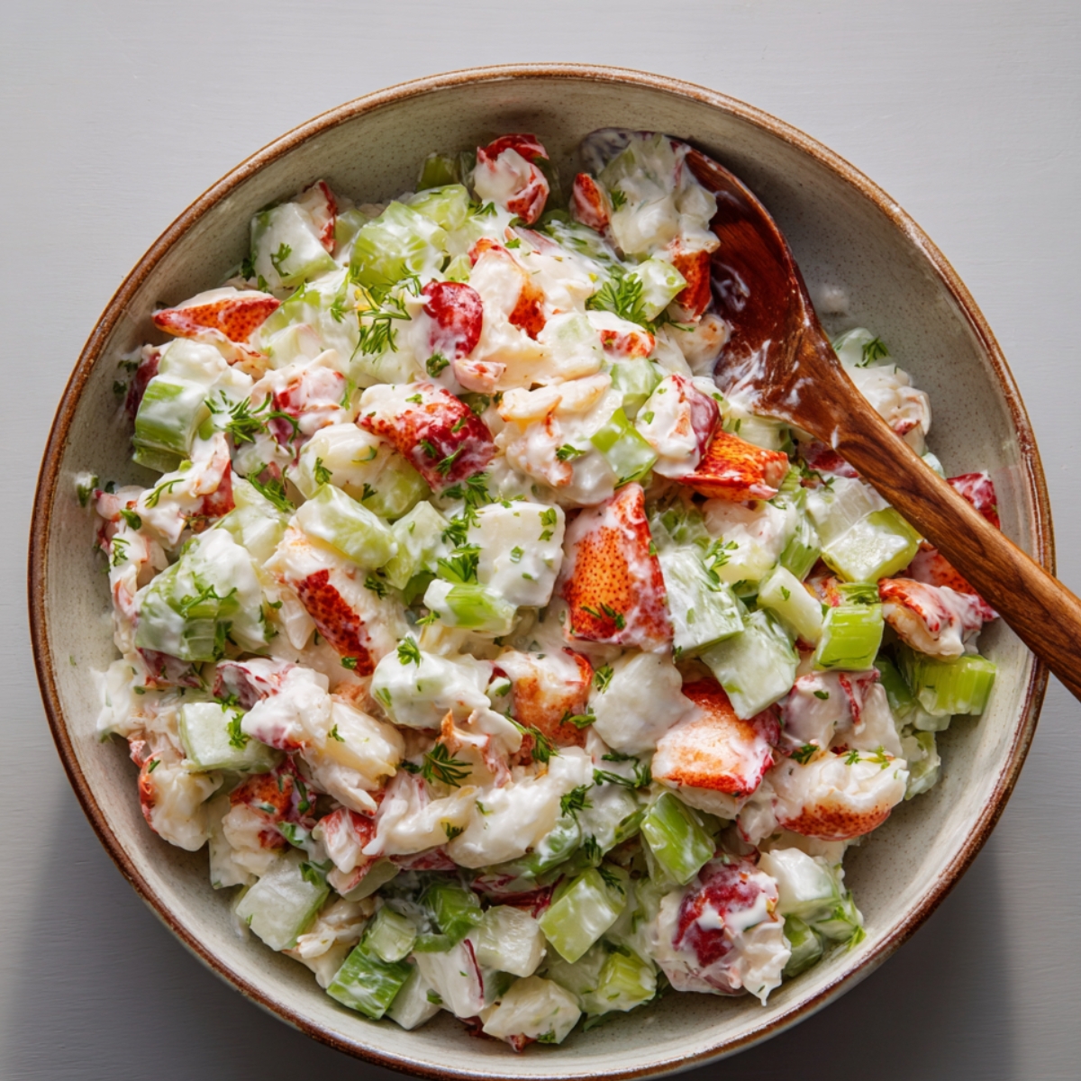 Lobster salad in a ceramic bowl with diced celery, lobster chunks, and creamy white dressing, garnished with chopped parsley and a wooden spoon.
