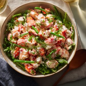 Bowl of creamy lobster salad with large lobster chunks, fresh dill, chopped scallions, and arugula, served in natural daylight with a wooden spoon beside the bowl.