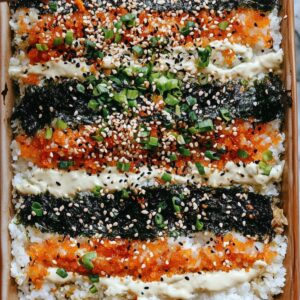 A rectangular sushi bake with neat horizontal layers of rice, orange tobiko (fish roe), nori seaweed, and mayonnaise drizzle. Garnished with sesame seeds and chopped green onions.