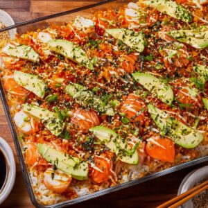 A glass baking dish filled with sushi bake, layered with white rice, avocado slices, raw salmon, imitation crab, and drizzled with sauces. Topped with chopped herbs, sesame seeds, and seaweed flakes for garnish.