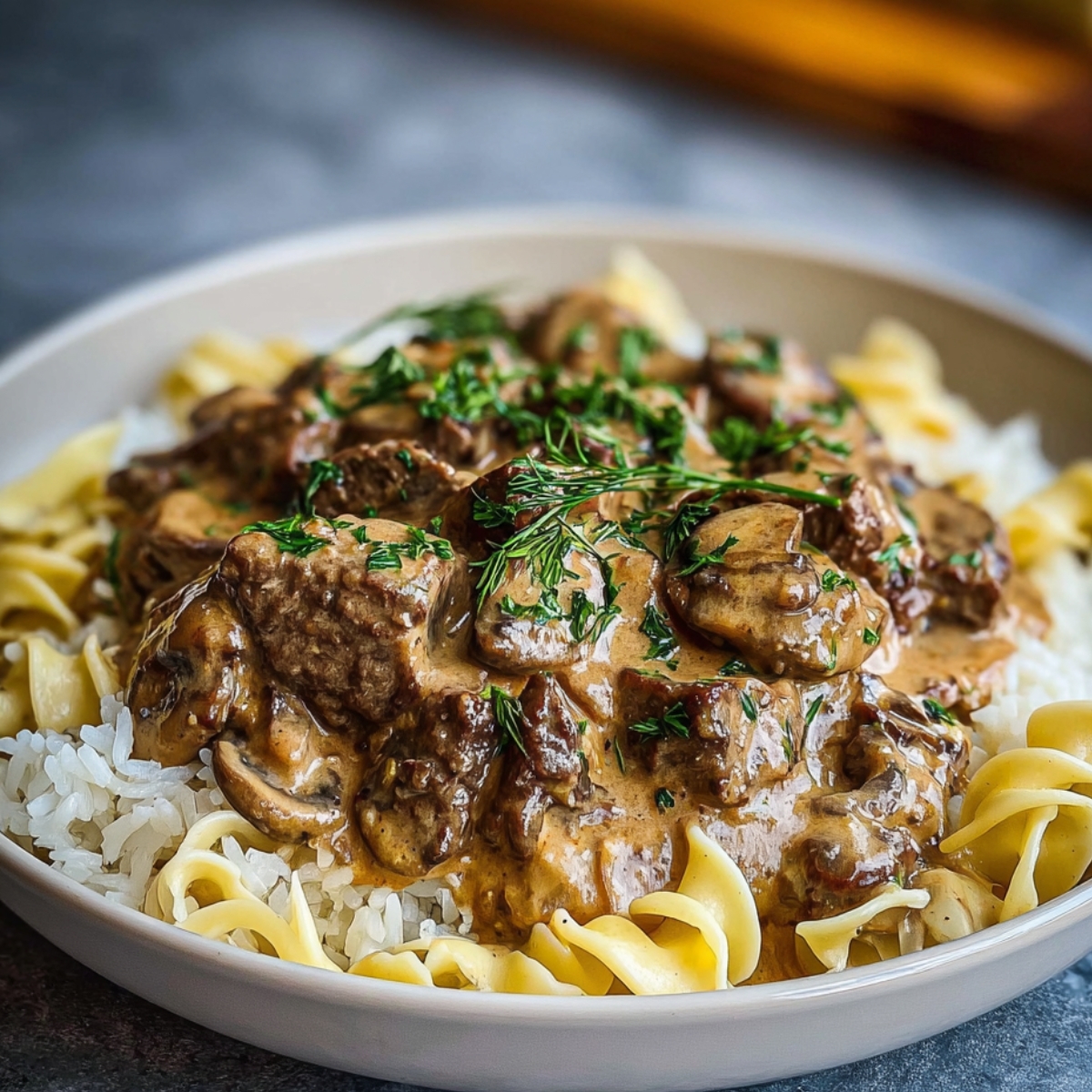 A bowl of homemade beef stroganoff with tender beef, mushrooms, and creamy sauce served over egg noodles and rice, garnished with fresh dill.