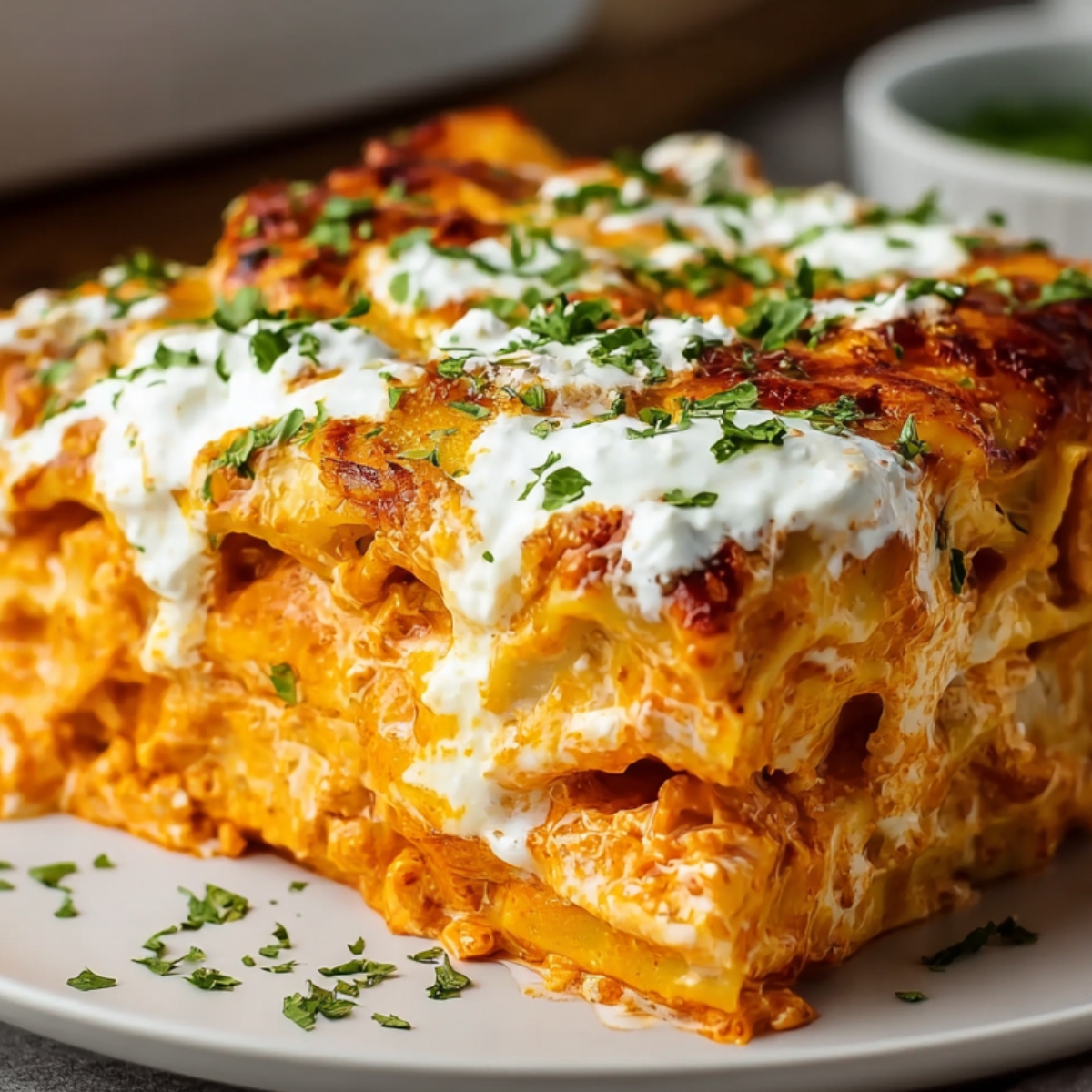 A slice of creamy buffalo chicken lasagna on a white plate, showing layers of pasta, orange sauce, melted cheese, and drizzled ranch with parsley garnish.