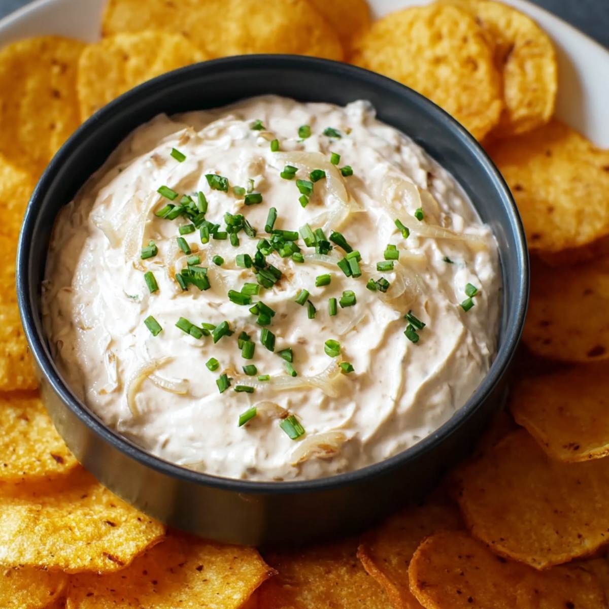 A bowl of onion dip recipe topped with caramelized onions and chives, surrounded by crispy chips.