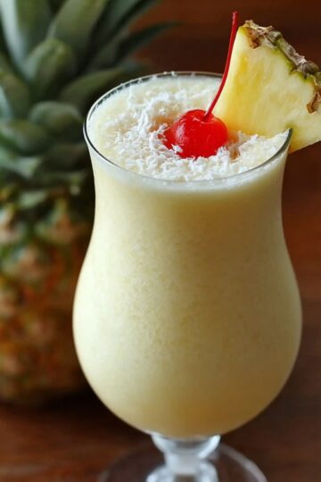 “Glass of homemade Pina Colada Recipe topped with shredded coconut, a cherry, and a pineapple wedge, with a fresh pineapple in the background.”