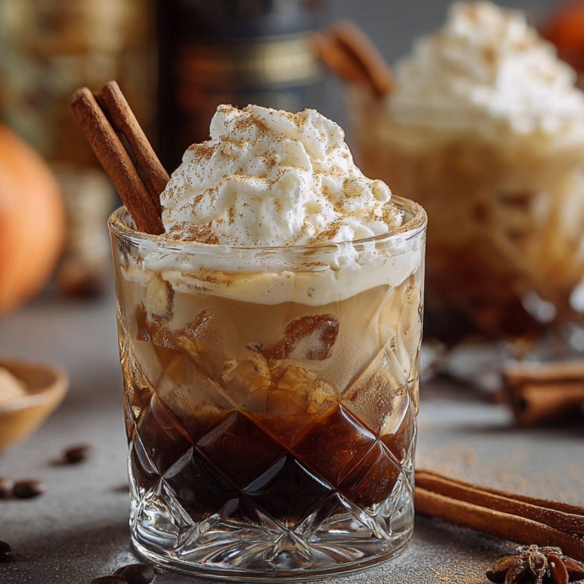 “A glass of Pumpkin Spice White Russian cocktail topped with whipped cream and sprinkled with cinnamon, garnished with two cinnamon sticks, sitting on a table with coffee beans and spices in the background.”