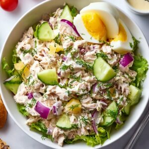 A bowl of Tuna Salad Recipe served on a bed of lettuce with chunks of cucumber, red onion, and pieces of hard-boiled egg, garnished with fresh dill.