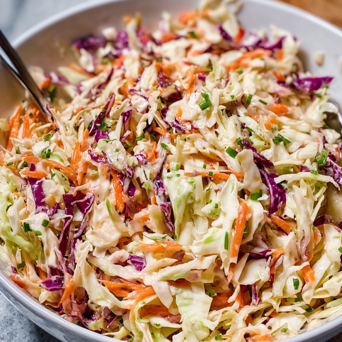A bowl of homemade creamy coleslaw recipe with shredded green and purple cabbage, carrots, and chopped chives, mixed with a mayonnaise-based dressing.
